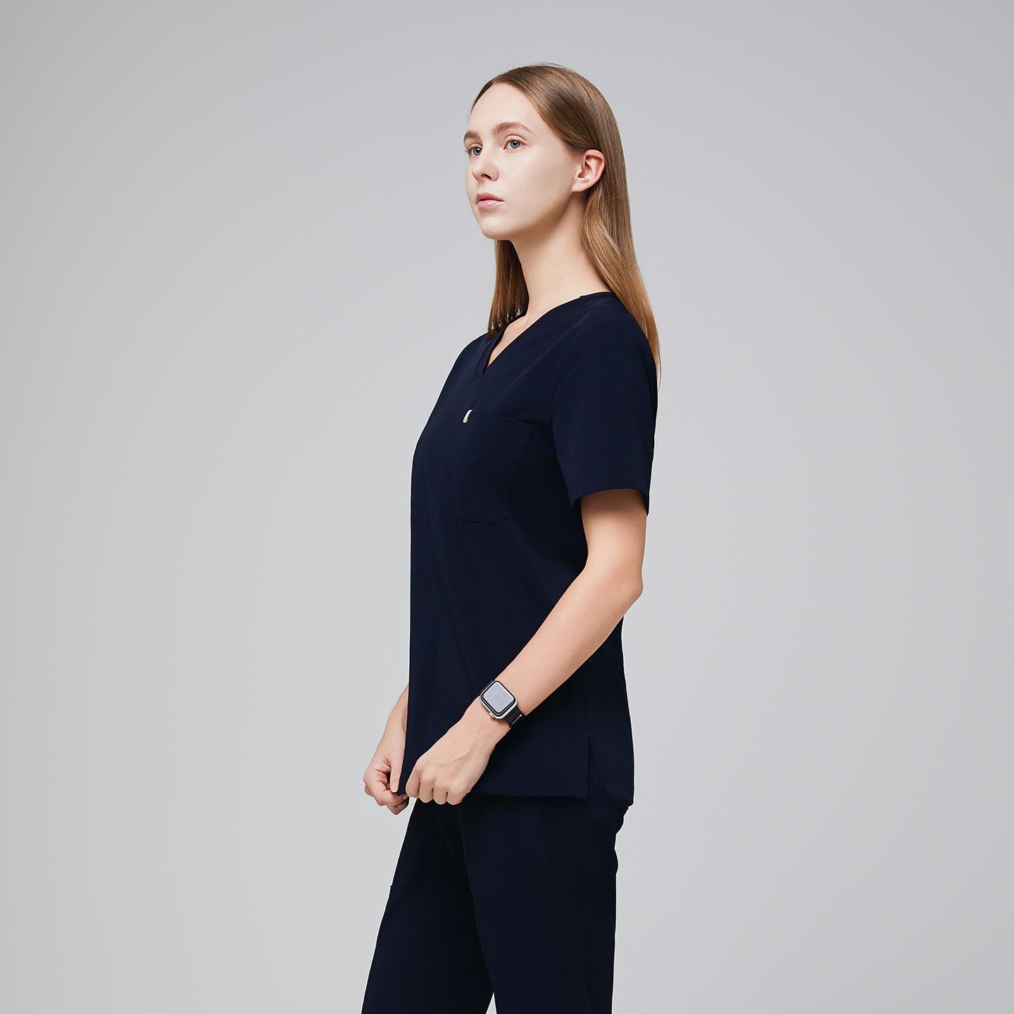 Image of Navy scrub top with single pocket paired with matching scrub pants, with the women holding her top and wearing a watch,Eco navy