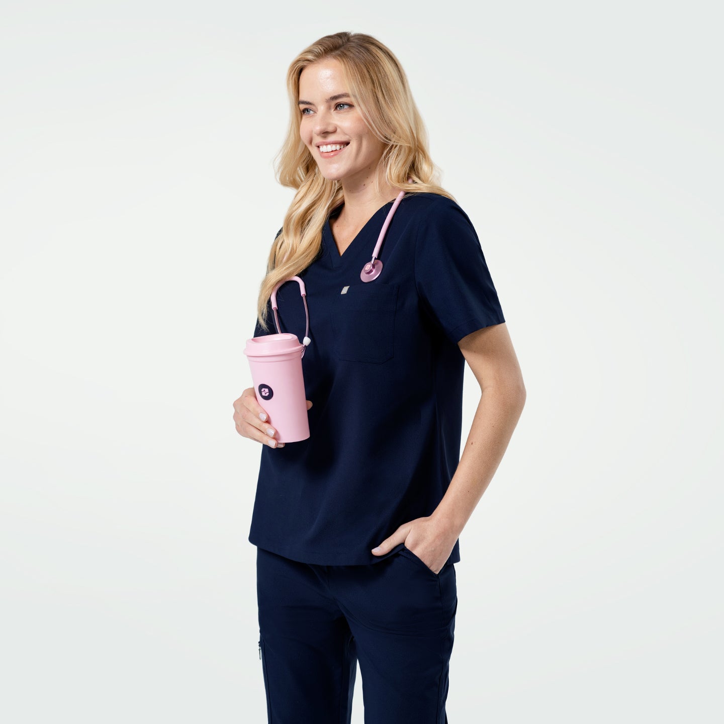 Image of The model is smiling while holding a pink tumbler in one hand, with a pink stethoscope draped around her neck, wearing a women’s Eco Navy single pocket scrub top.