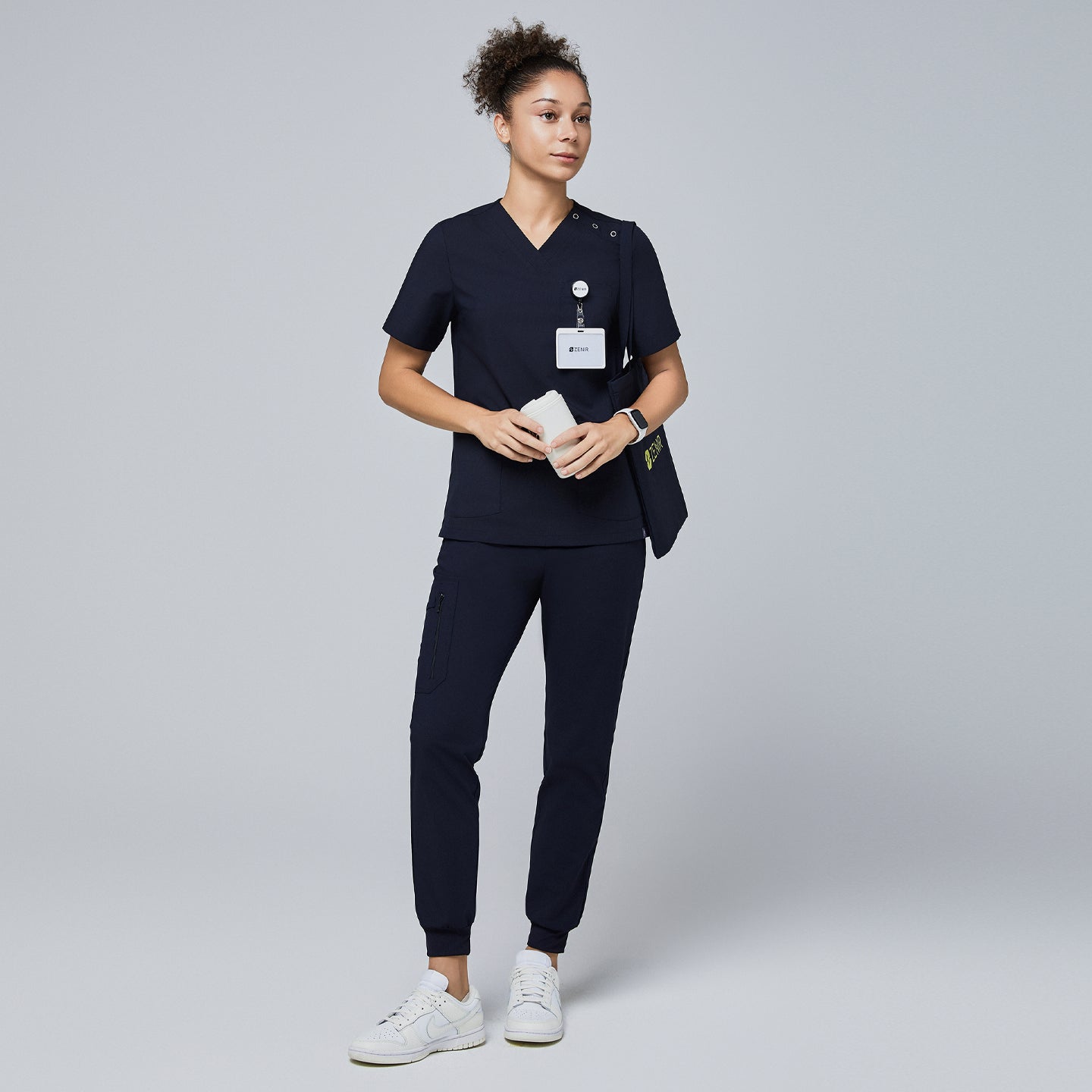 Image of Female model wearing a navy V-neck scrub top with shoulder buttons, navy jogger scrub pants, a name badge, and a Zenir tote bag, holding a reusable cup,Eco Navy
