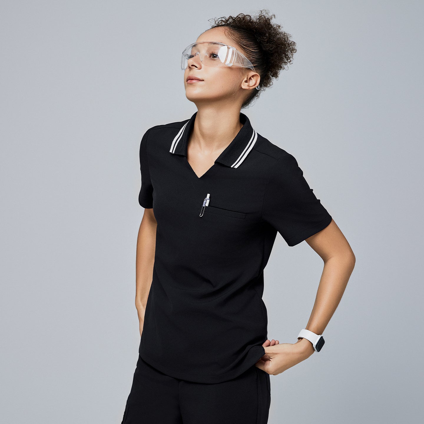 Image of Female model wearing a jet black essential polo scrub top with a striped collar, looking upward, accessorized with safety glasses and a smartwatch,Jet Black