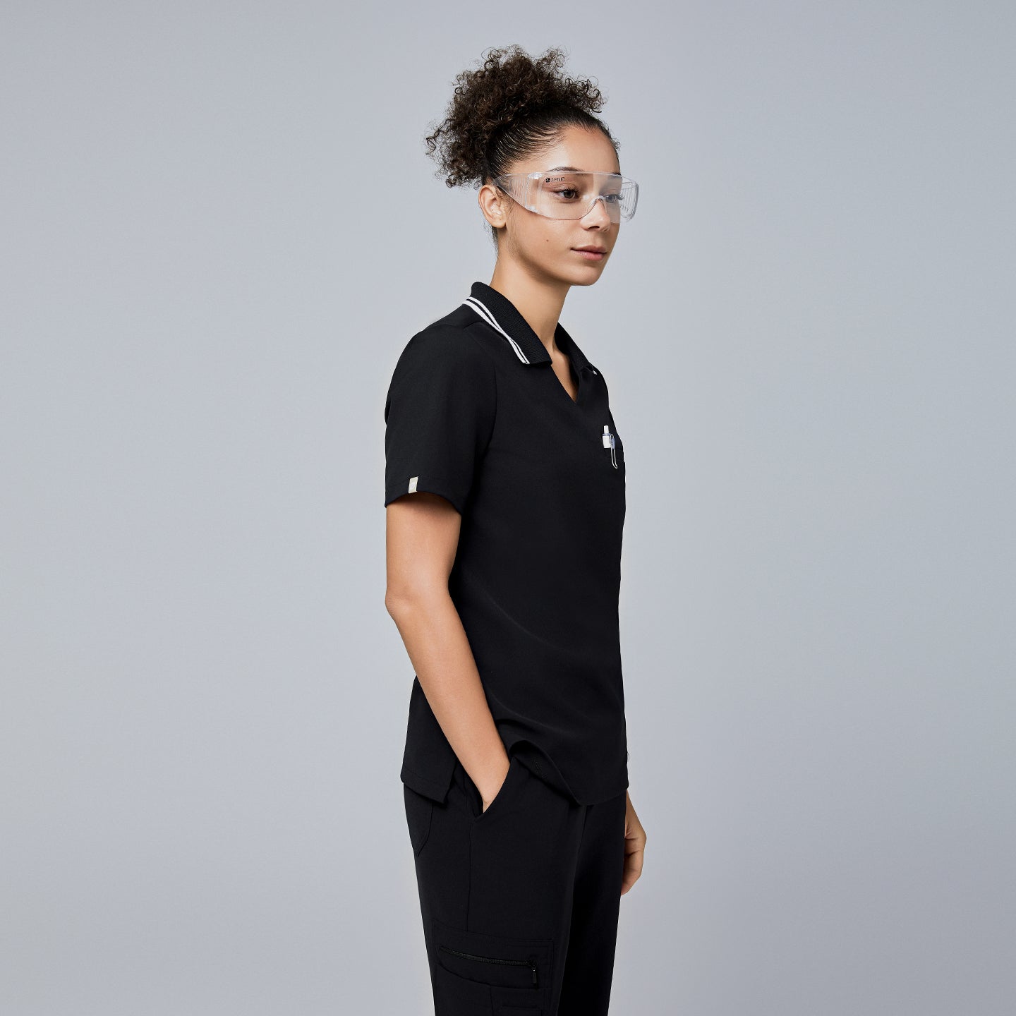 Image of Female model wearing a jet black essential polo scrub top with a striped collar, viewed from the side, accessorized with safety glasses and her hands in her pockets,Jet Black