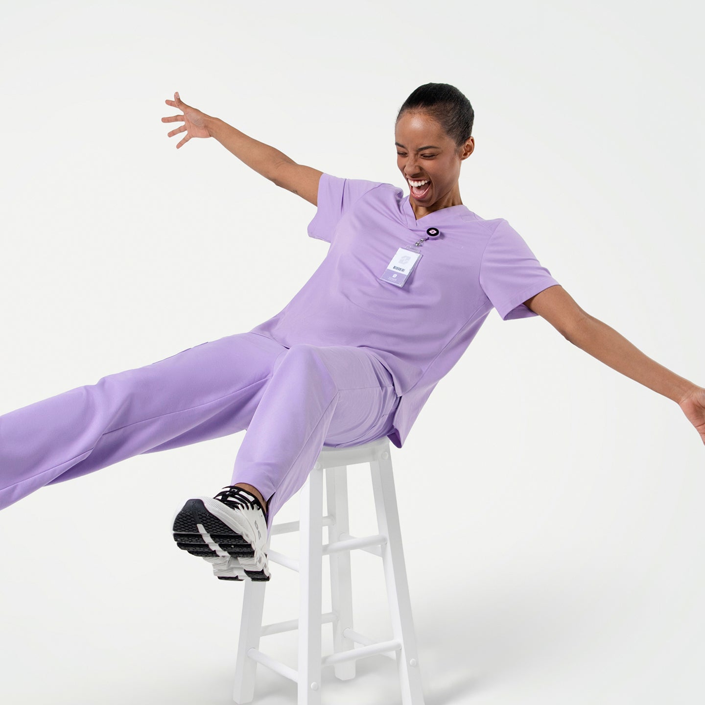 Image of Energetic pose in the Women's Lavender Pleated Shoulder Scrub Top and pants, sitting on a stool with arms wide open, full body shot