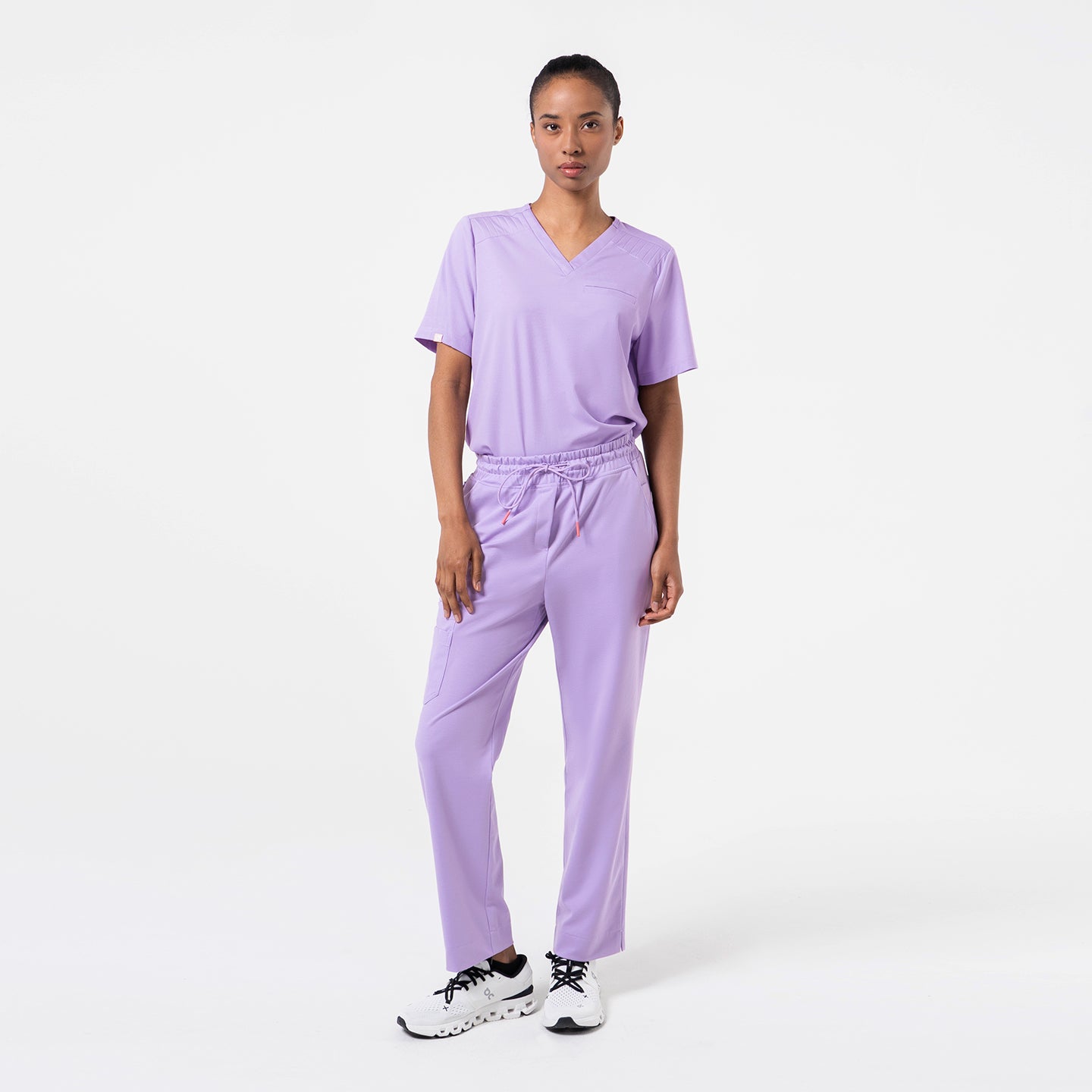 Image of A model wears a lavender pleated-shoulder scrub top with a clean V-neck and subtle pleat detailing along the shoulders. The lightweight lavender fabric drapes smoothly, offering comfort and ease of movement.