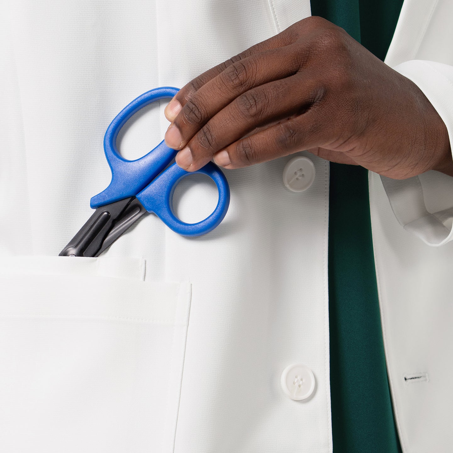 Image of A close-up view of the LCM-201 lab coat highlights its large, practical front pocket as a hand places a pair of blue medical shears inside, emphasizing the coat’s functional storage space, clean stitching, and professional, durable white fabric designed for everyday clinical use.