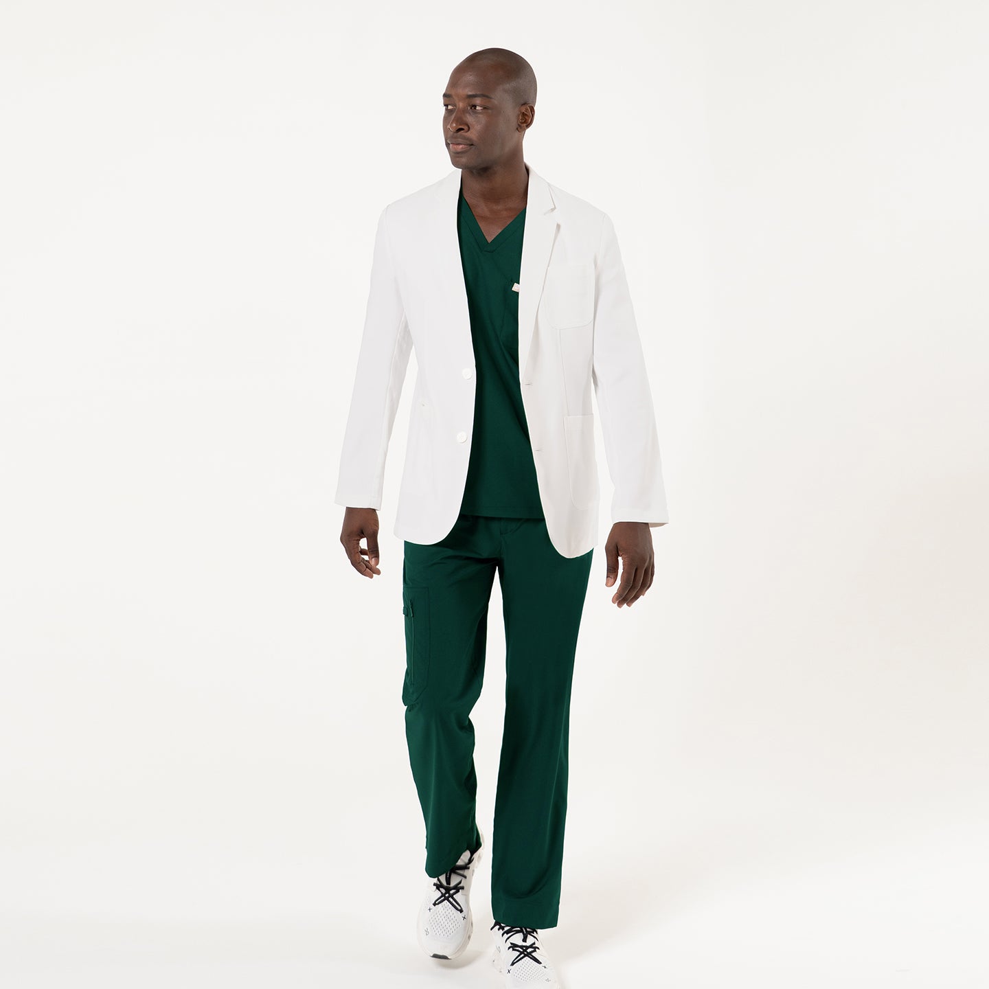 Image of A model wearing a crisp white LCM-201 lab coat stands confidently with a relaxed, open stance, showcasing the coat’s structured tailoring, clean notched lapels, spacious front pockets, and smooth, professional drape that pairs seamlessly with the black scrub set underneath for a polished clinical look.