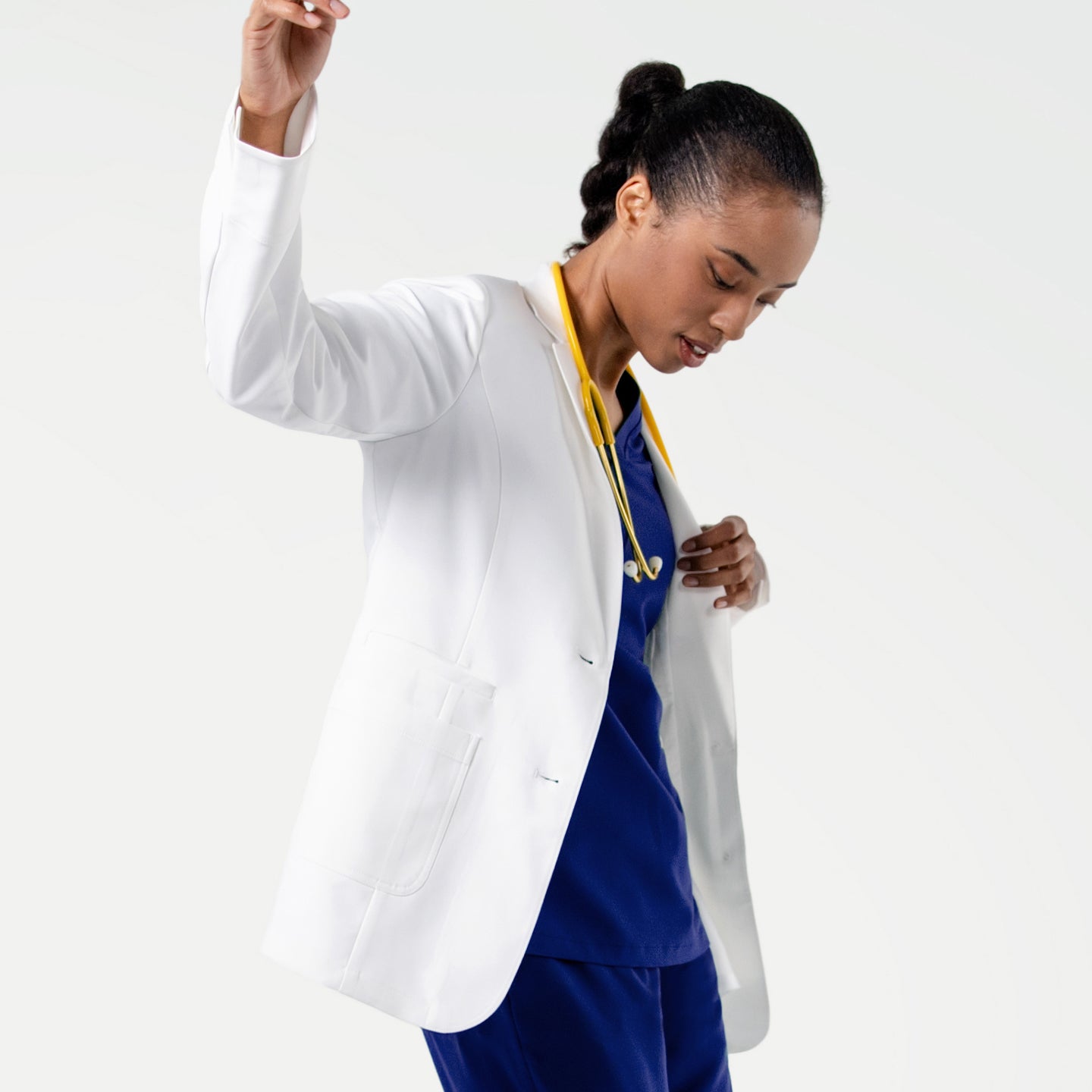 Image of Showcasing the soft stretch and refined structure of the LCW-202 lab coat, the model moves with ease as the crisp white fabric flows over her blue scrubs, complemented by a yellow stethoscope resting at her neckline.