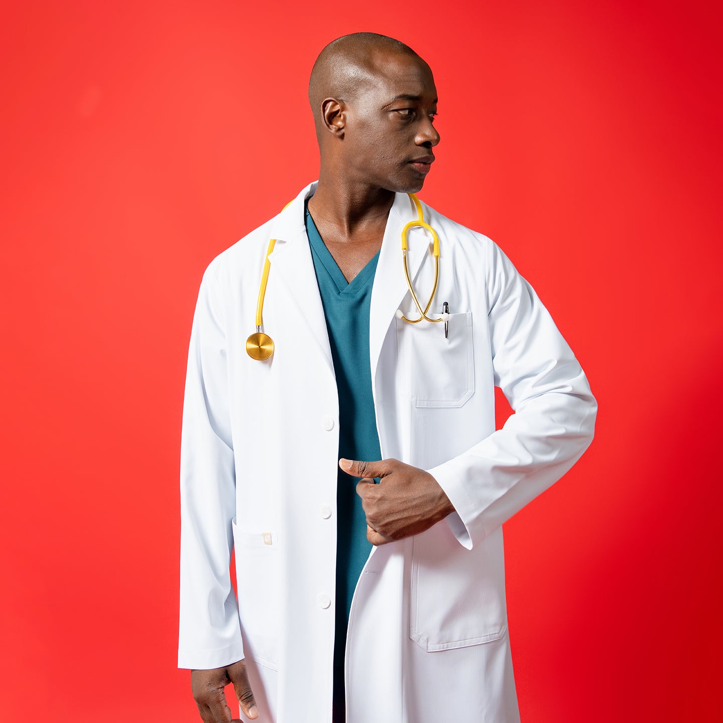 Image of A men’s short lab coat LCM-02 in white is worn over teal scrubs, featuring a chest pocket and lower patch pockets. The model, posed against a solid red background, wears a yellow stethoscope around the neck and adjusts the front of the coat while looking to the side.