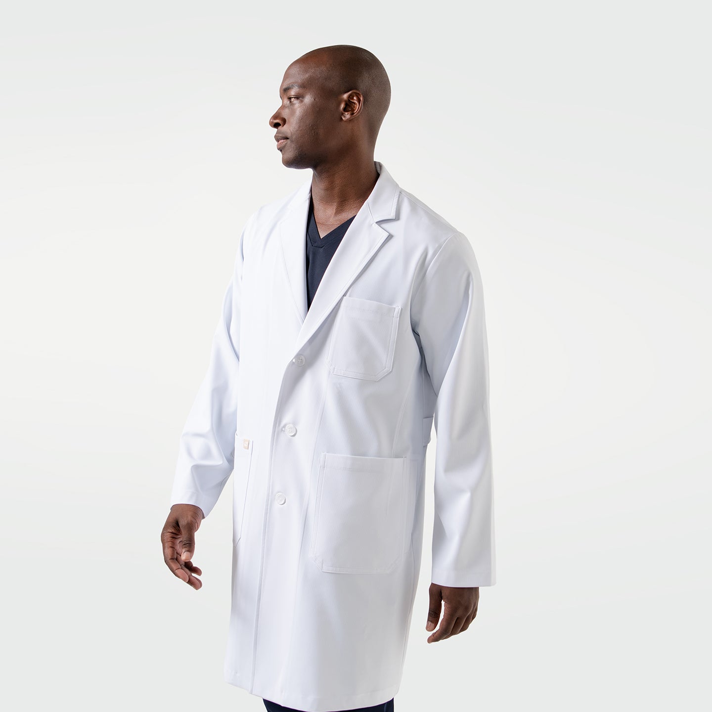 Image of The male model wears the Men’s LCM-02 White Lab Coat over navy scrubs, standing in a side-facing pose with a composed and professional expression.