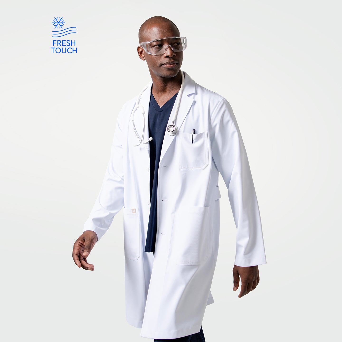 Image of Male model wearing the Men’s LCM-02 White Lab Coat over navy scrubs, accessorized with a stethoscope and clear protective glasses, walking with a confident stride.
