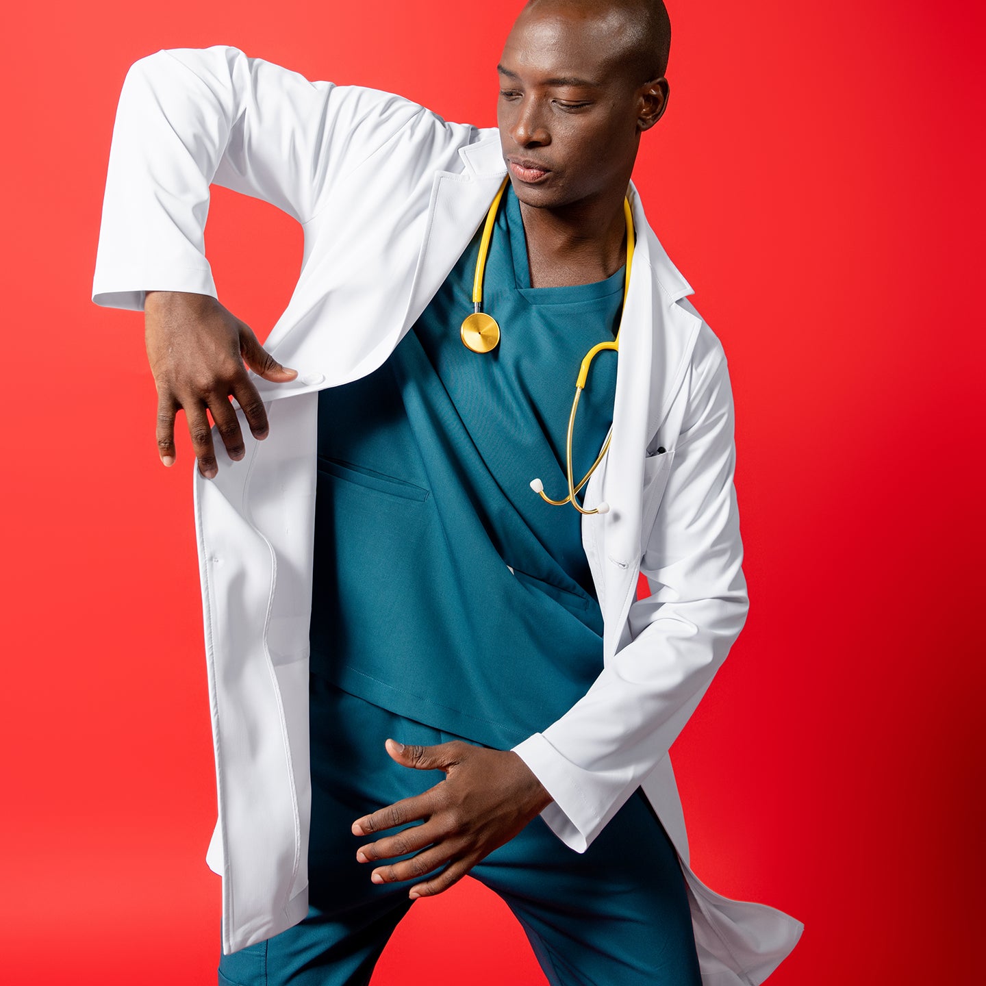 Image of LCM-02 in white is styled over teal scrubs. The model strikes a dynamic pose against a solid red background, with a yellow stethoscope around the neck and one arm lifted to adjust the coat.