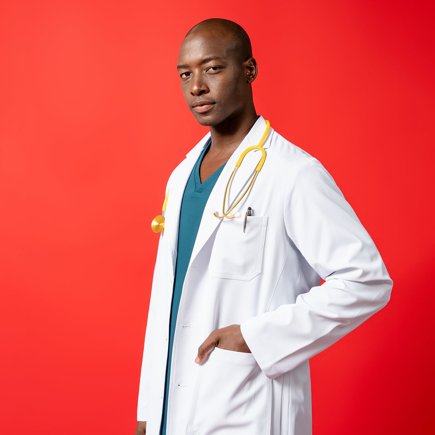 Image of The LCM-02 Lab coat features a tailored fit with a chest pocket and lower patch pockets, styled with a yellow stethoscope draped around the neck. Model poses against a bold red background with one hand in the pocket.