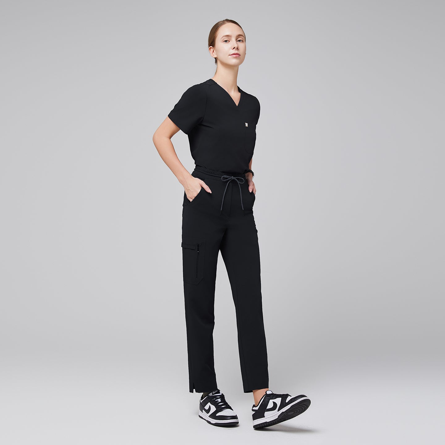 Image of Model wearing a scrub set featuring a V-neck top and drawstring pants with zipper pockets and split hem, paired with black and white sneakers for a modern, professional style,Midnight Black