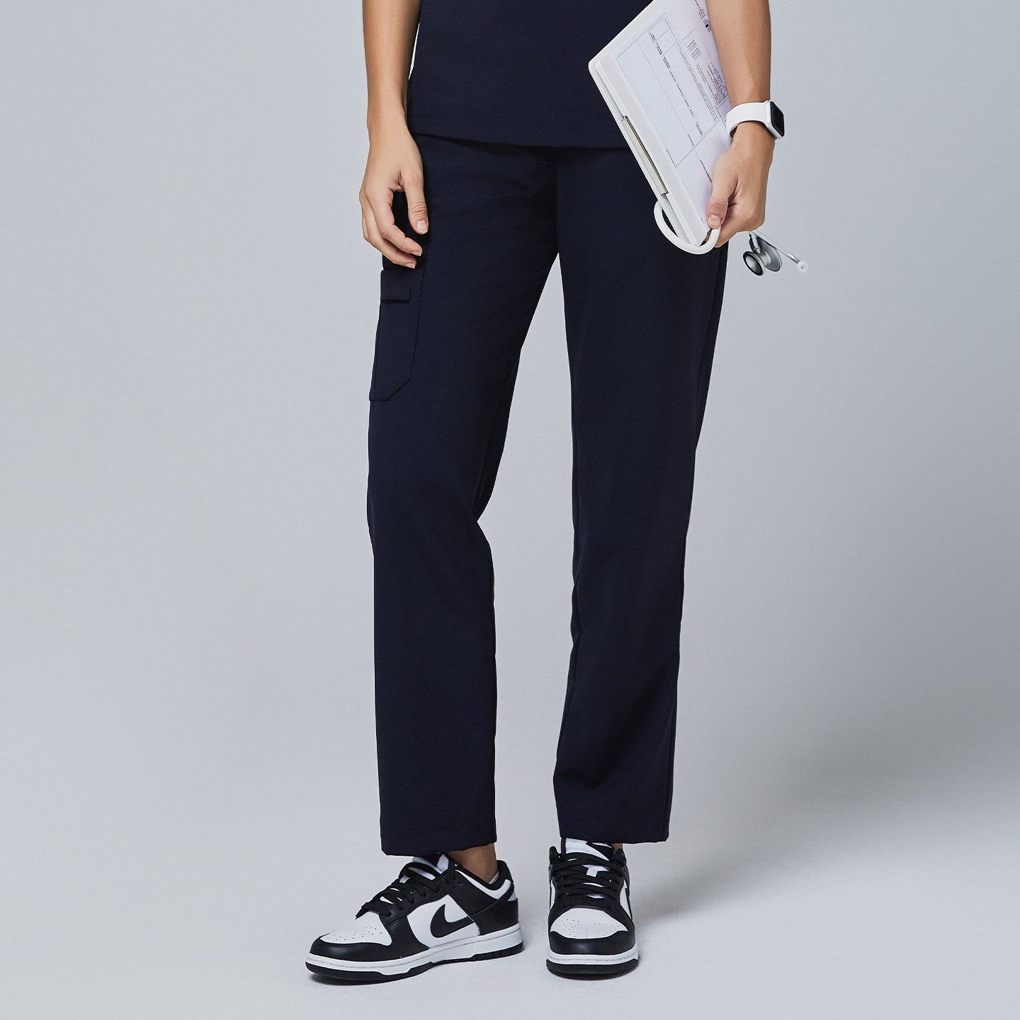Image of Navy utility scrub pants featuring a side pocket, worn with a navy top and paired with black-and-white sneakers, holding a clipboard and stethoscope,Midnight Navy