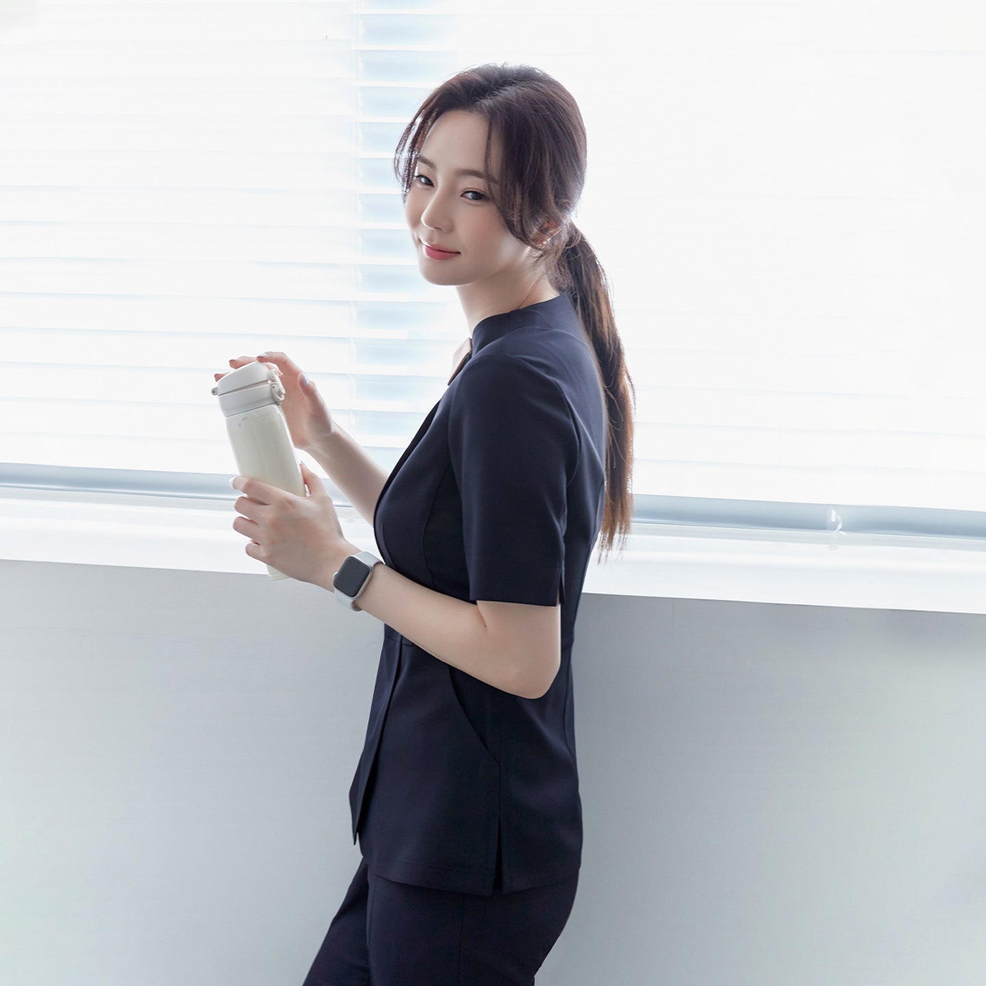Image of A woman in a navy round-neck front zipper top and matching pants, standing by a window, holding a white tumbler, and wearing a smartwatch,Mir Navy