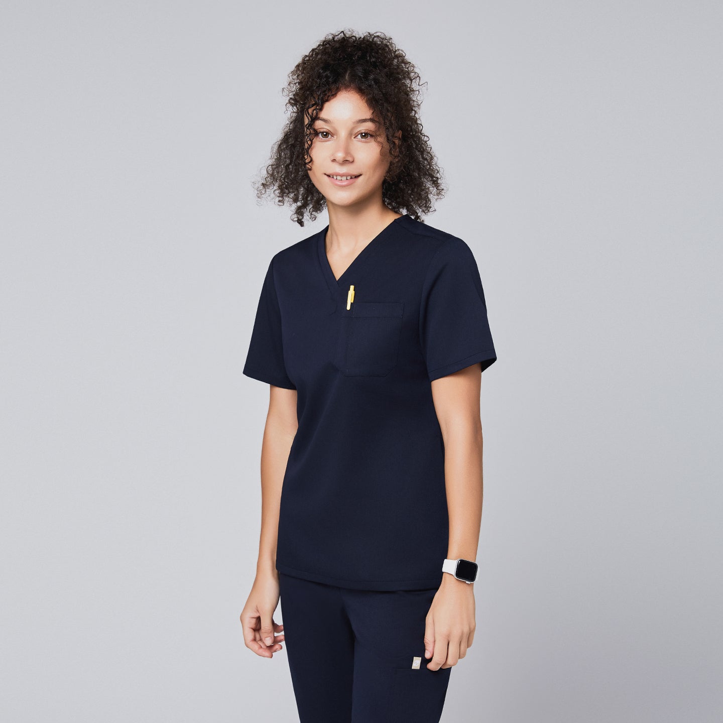 Image of Woman in a navy scrub top with a chest pocket holding a yellow pen, smiling,Navy