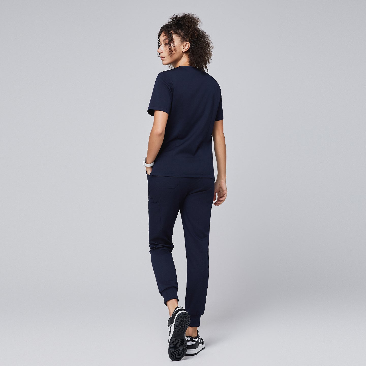 Image of Woman wearing a navy scrub set featuring a short-sleeve top and jogger-style pants, shown from the back, with hands in pockets and black and white sneakers,Navy