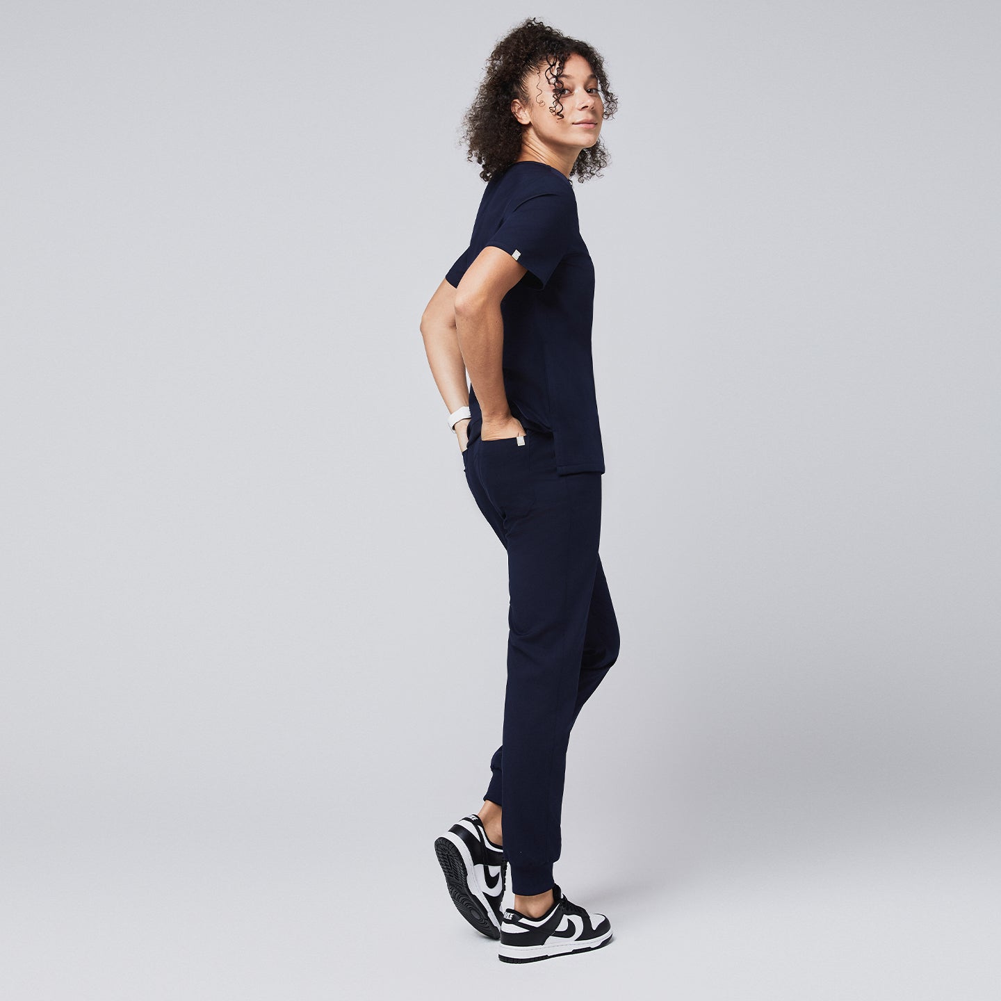 Image of Woman wearing a navy scrub top with a chest pocket and jogger-style scrub pants, standing in a side profile pose with hands in pockets, paired with black and white sneakers,Navy