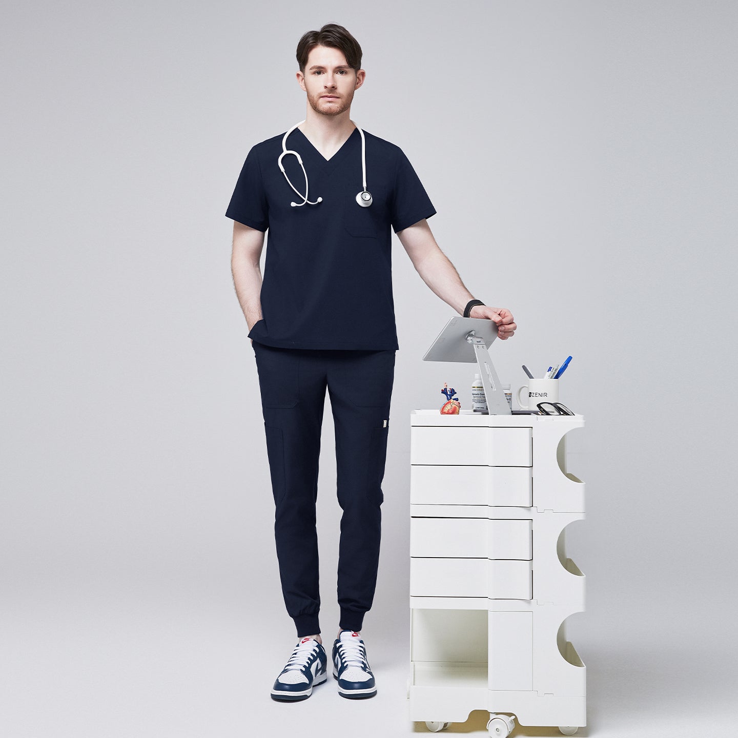 Image of Man wearing navy V-neck scrub top and 8-pocket jogger scrub pants, accessorized with a stethoscope, standing beside a white medical cart,Navy