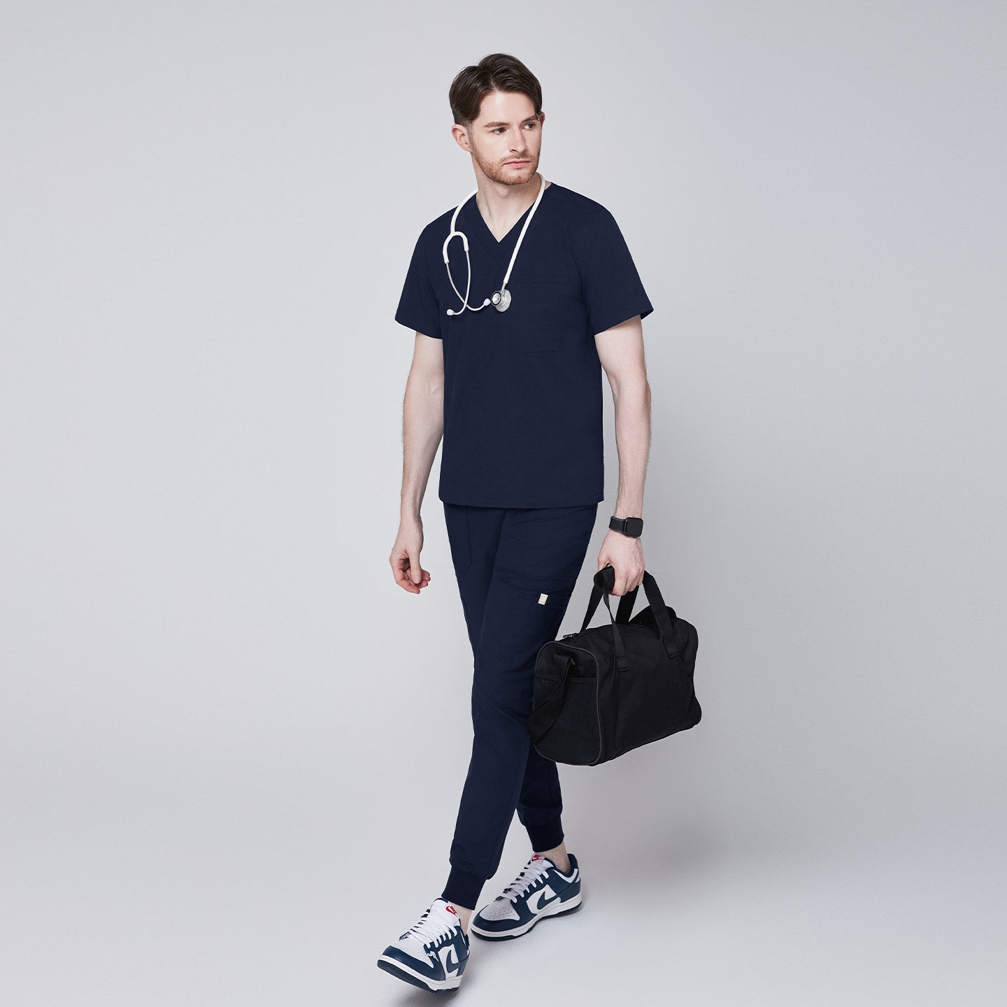 Image of Man wearing navy V-neck scrub top and jogger scrub pants with a stethoscope and black duffle bag, paired with navy sneakers,Navy