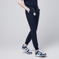 Image of Side angle of navy jogger scrub pants with 8 pockets, drawstring waistband, and cuffed ankles, paired with navy sneakers and a name badge,Navy