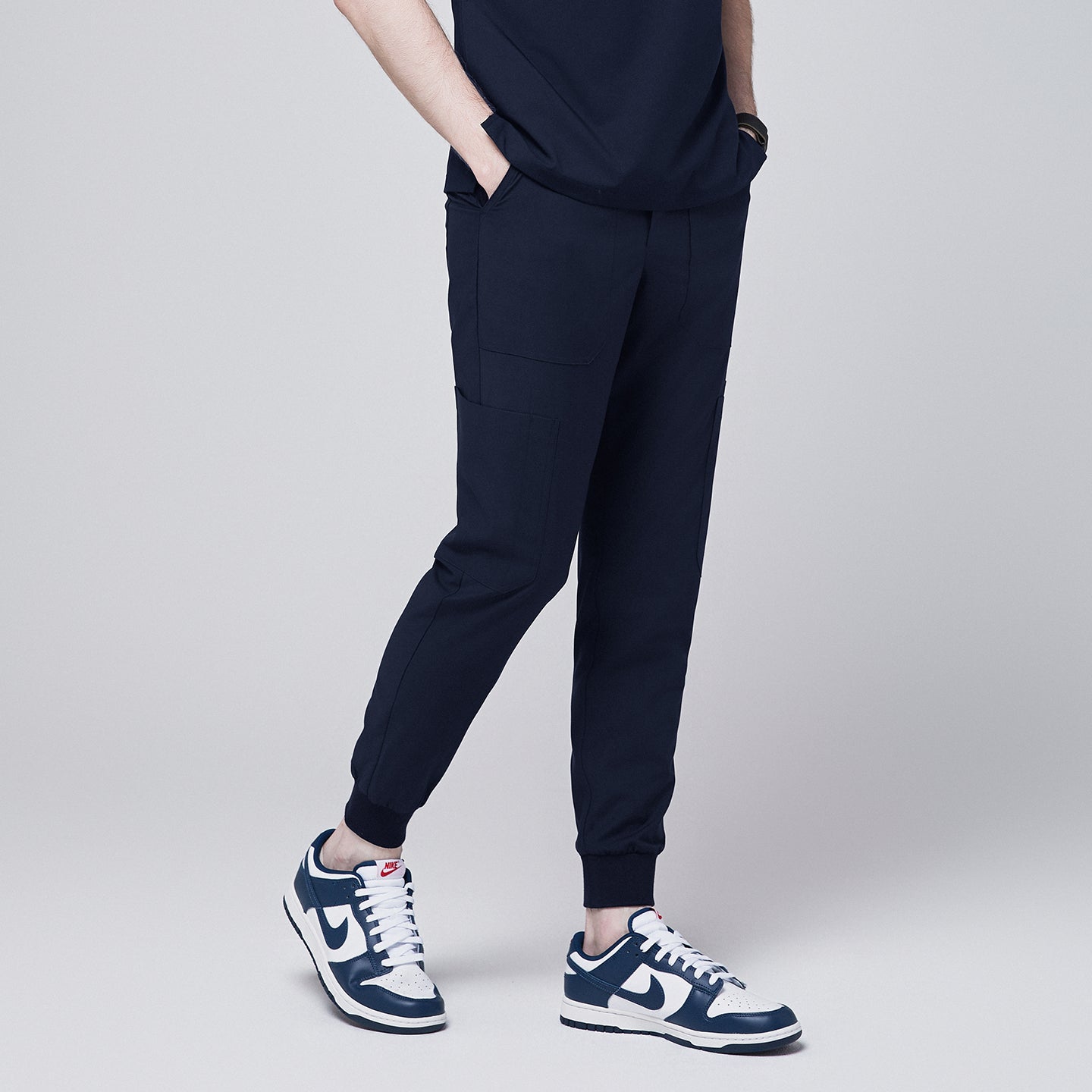 Side view of navy jogger scrub pants with 8 pockets, cuffed ankles, and hands in pockets, paired with navy sneakers,Navy