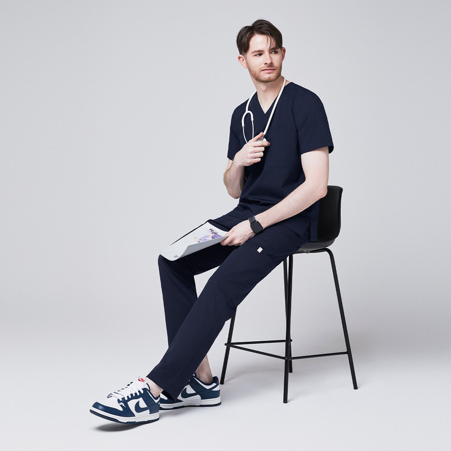 Image of man seated, wearing Zenir navy 8-pocket scrub pants, matching top, stethoscope, and blue sneakers, holding a clipboard,Navy