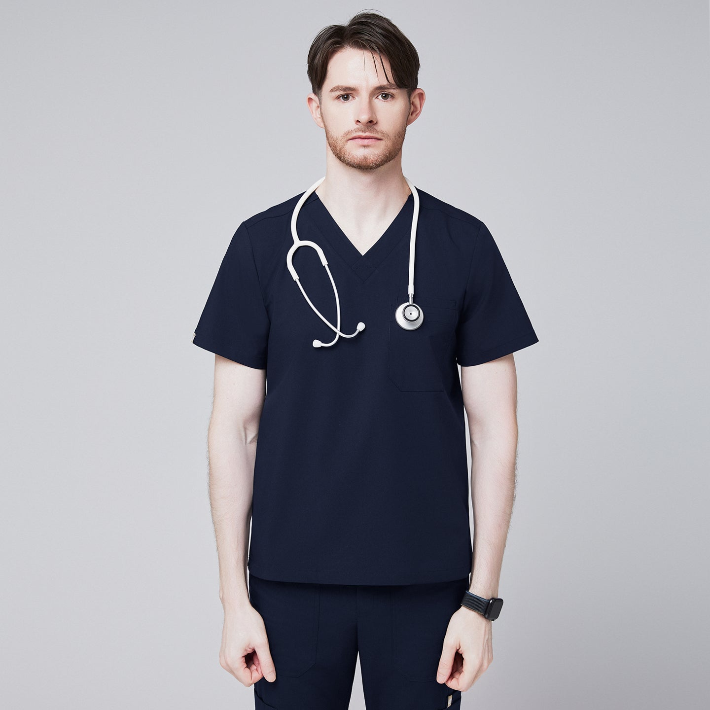 Image of Male healthcare professional wearing a navy V-neck, three-pocket scrub top with stethoscope around neck, standing against a plain background,Navy