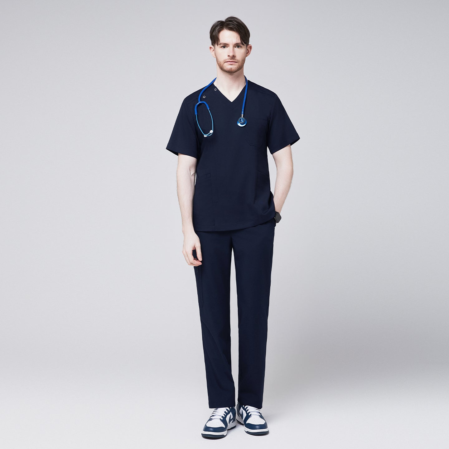 Image of Full-body view of a man wearing a navy scrub top with a front zipper and matching pants, accessorized with a blue stethoscope and casual sneakers,Navy