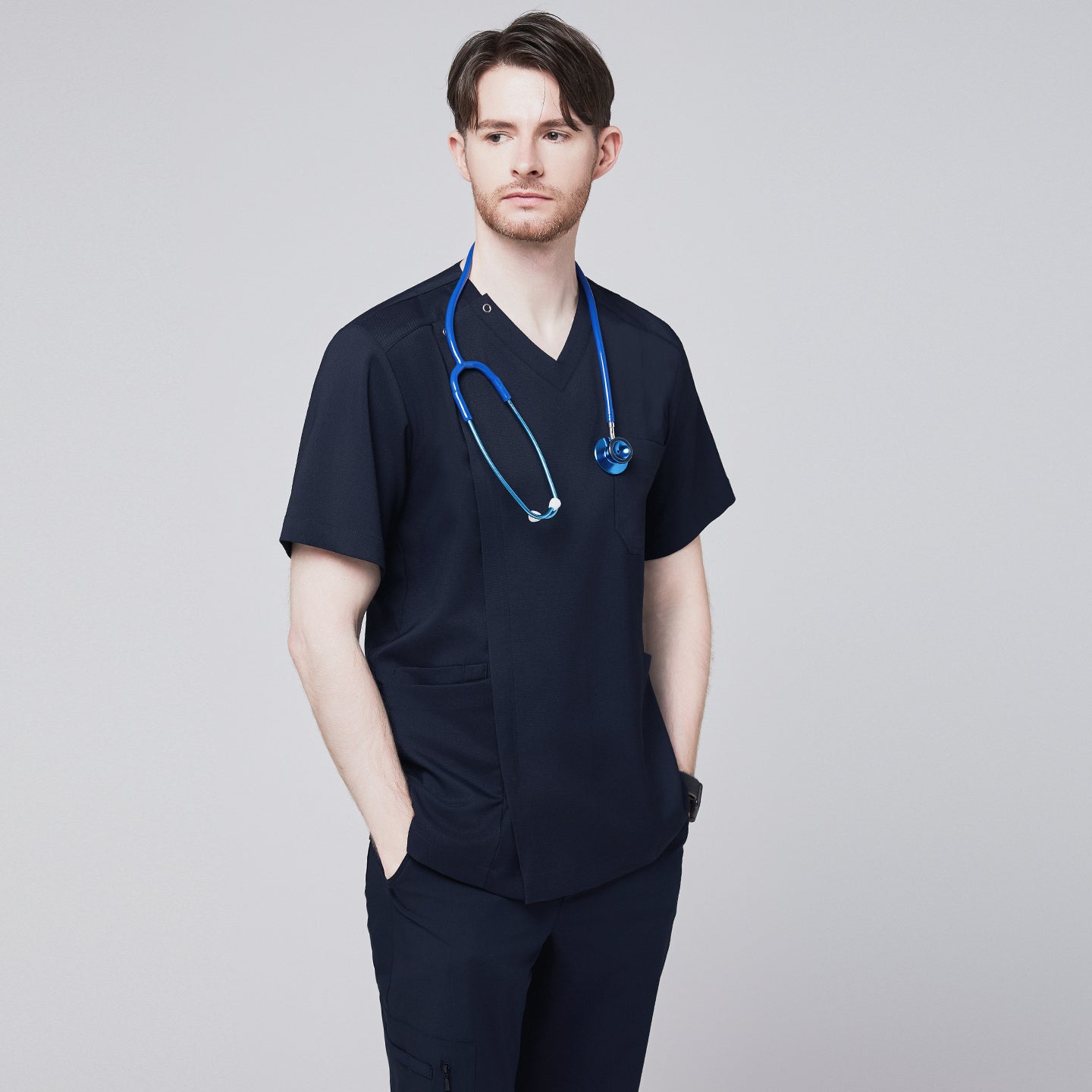 Image of Man wearing a navy scrub top with a front zipper detail, hands in pockets, styled with a stethoscope around his neck, looking confidently to the side,Navy