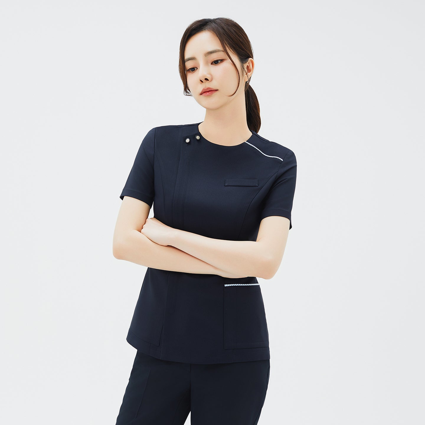 Image of Woman crossing her arms in a navy scrub top featuring snap-button accents near the neckline and white piping around the pockets,Navy
