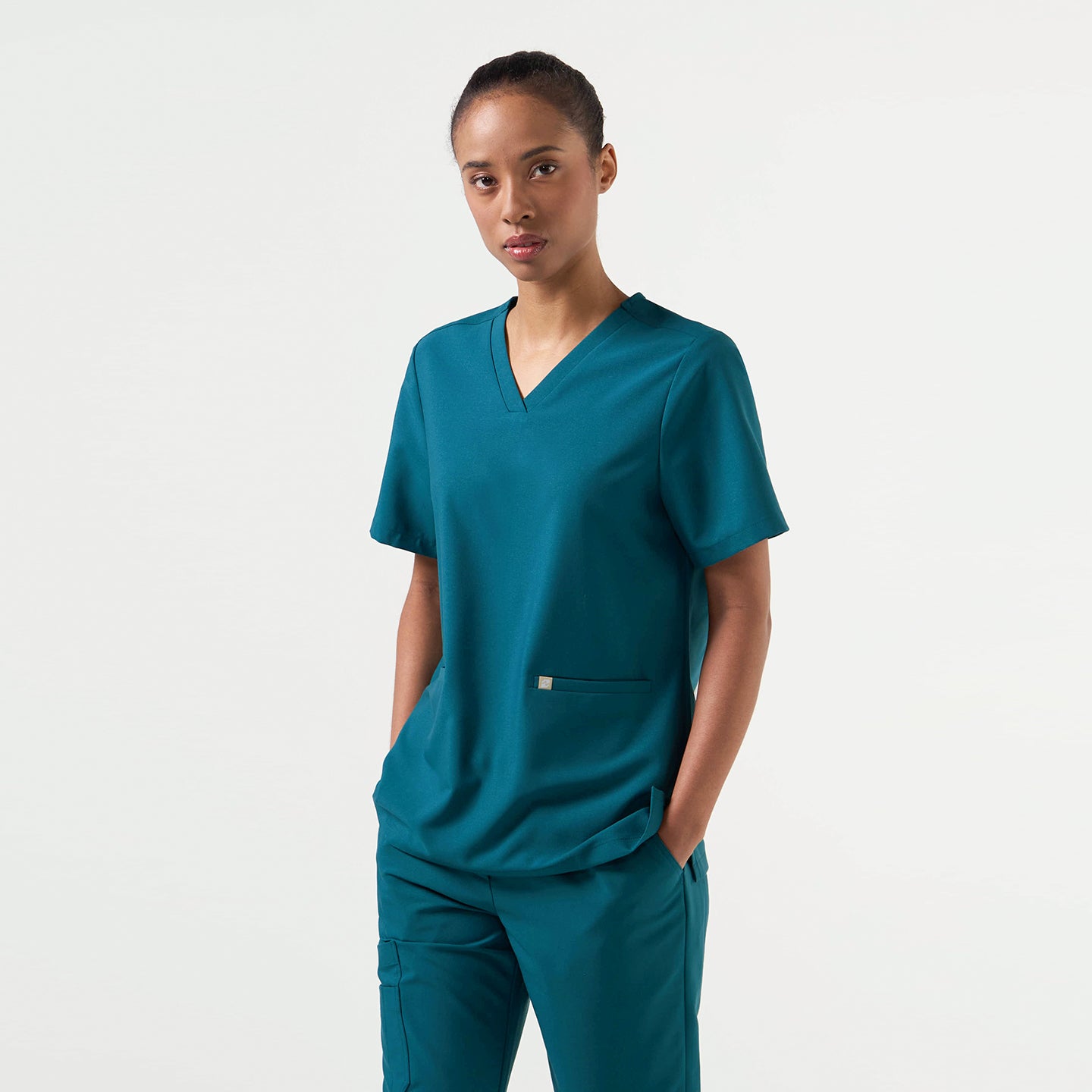 Image of Standing in the Women's Ocean Teal Double Pocket Scrub Top and pants, facing forward with hands in pockets, upper body view