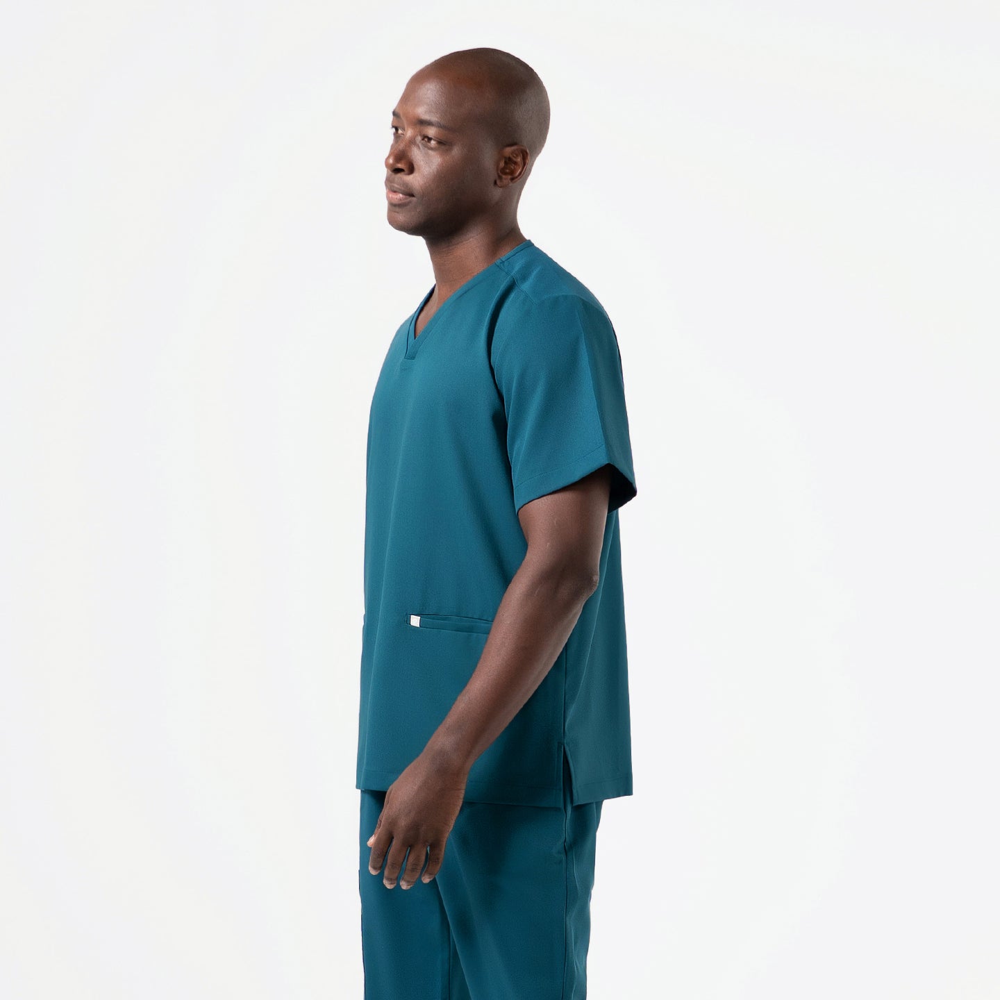 Side-profile view of a model wearing a Pine-Green double-pocket scrub top, showing the relaxed V-neck design, short sleeves, and a straight, easy-fit silhouette, with one hand resting naturally at his side to highlight the top’s smooth, flexible fabric and minimal utility pocket.