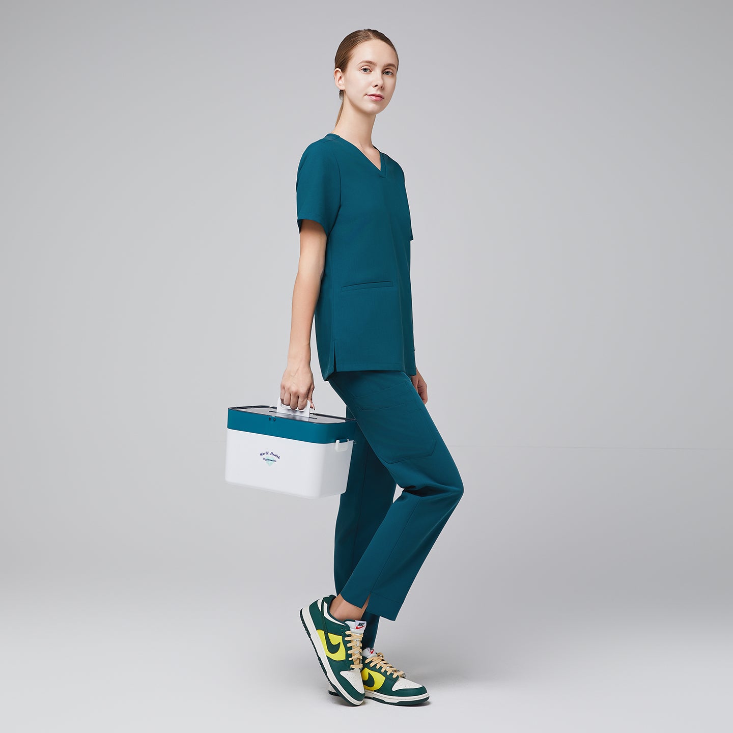 Full body shot with ocean teal scrub top and ocean teal straight scrub pants with a split hem, with green sneakers and a medical box in hand,Ocean teal