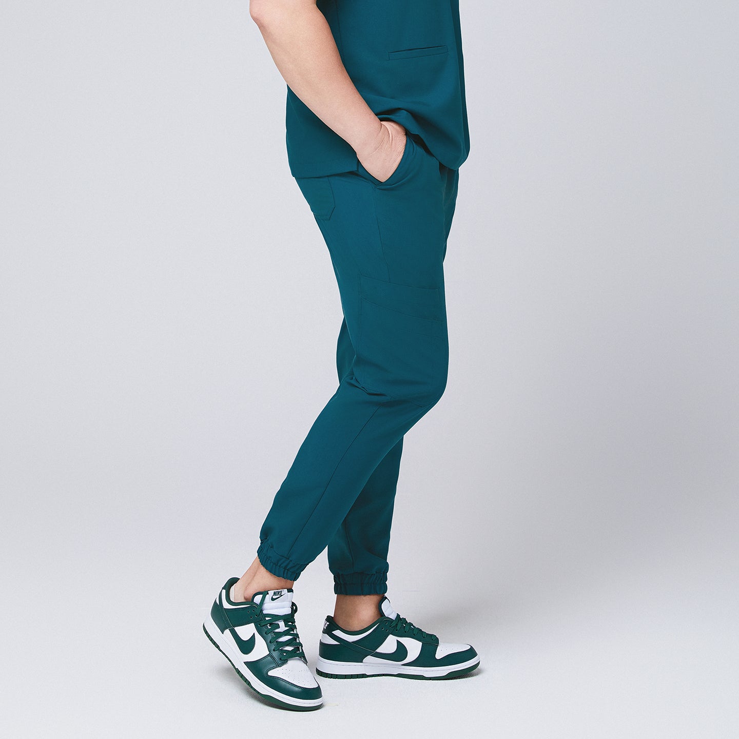 Image of Ocean teal jogger scrub pants made of stretchy fabric and matching ocean teal scrub top, with teal sneakers and hands in pocket,Ocean teal