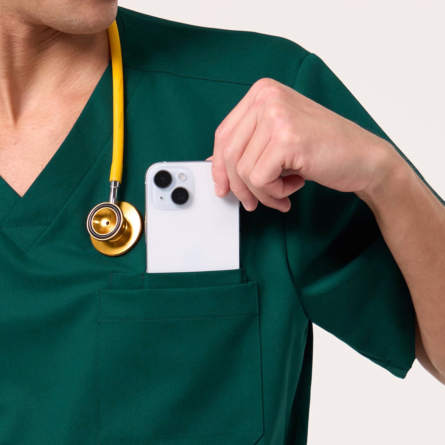 Image of Wearing a Pine Green scrub top, the male model places a smartphone into the chest pocket while a yellow stethoscope hangs around his neck.