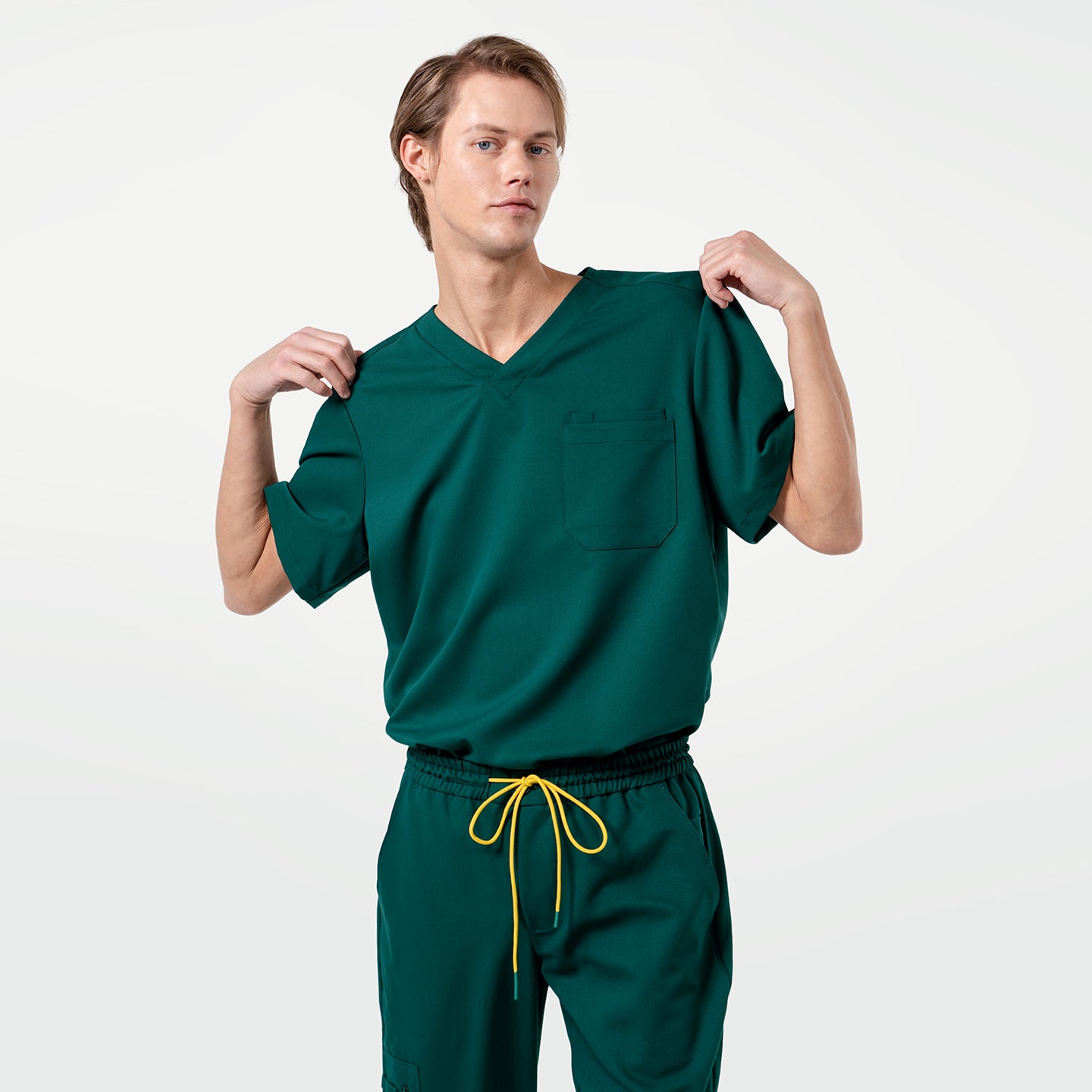 Image of A male model wearing an Eco Hunter Green scrub top and pants, posing with both hands lifting the shirt slightly at the shoulders.