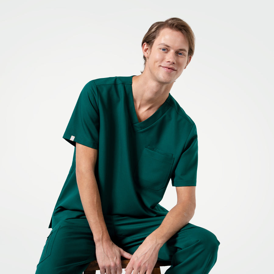 Wearing a Pine Green 3 Pocket Scrub Top, the model sits comfortably with a slight smile.