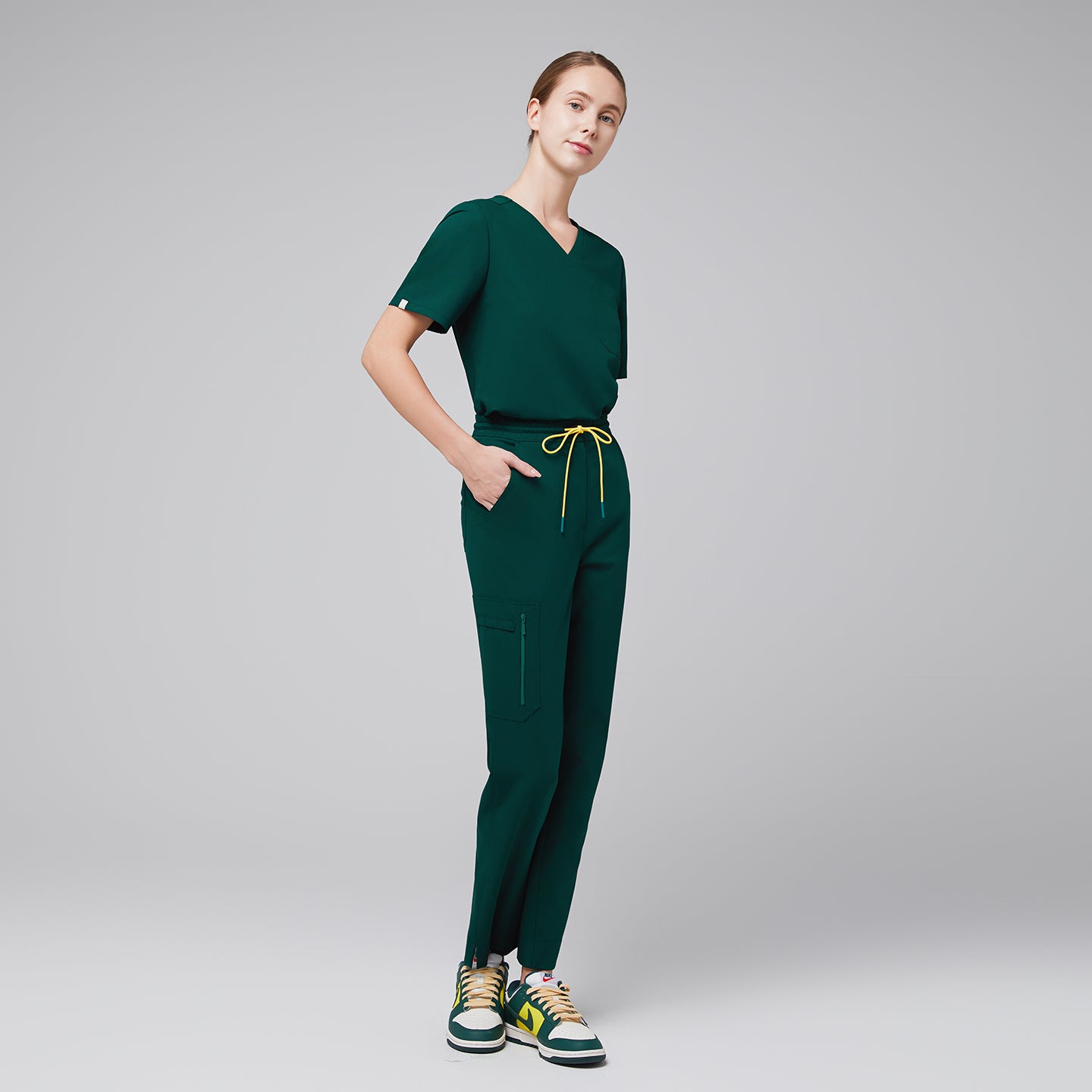 Image of A woman dressed in a pine green scrub set, featuring a relaxed V-neck top with three pockets and tapered pants with yellow drawstring accents, styled with sporty sneakers,Pine Green