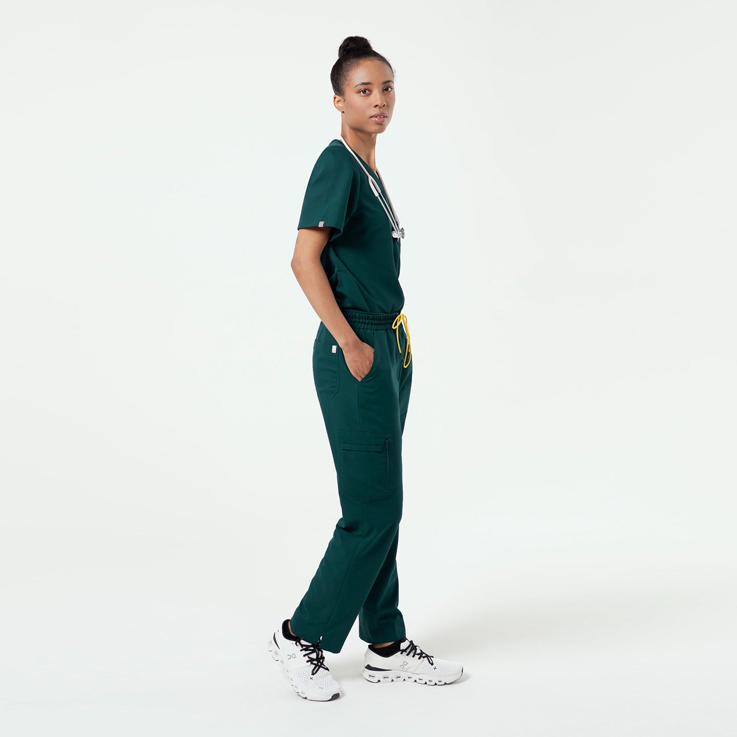 Model wearing a Pine Green 3-Pocket Scrub Top with matching jogger scrub pants, posing in a side profile with hands in pockets against a clean studio background.