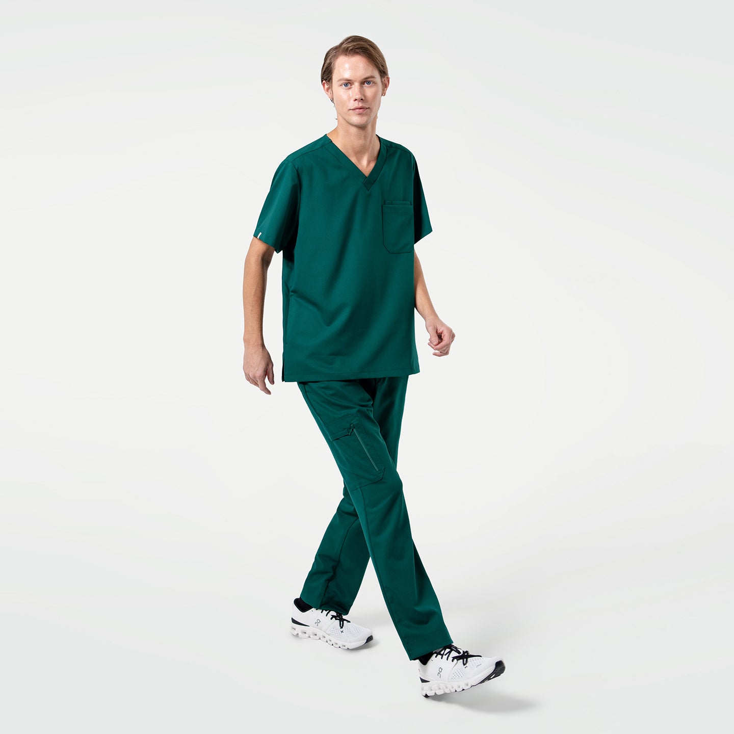 Image of A male model is shown wearing Pine-Green 3-Pocket Scrub Top, styled with matching Pine-Green scrub pants for a clean and coordinated medical uniform look. The scrub top features a classic V-neckline and a practical chest pocket, offering both functionality and a streamlined appearance suitable for clinical environments. The model is captured mid-step, highlighting the comfortable fit and easy mobility of the fabric, making it ideal for long, active shifts in healthcare settings.