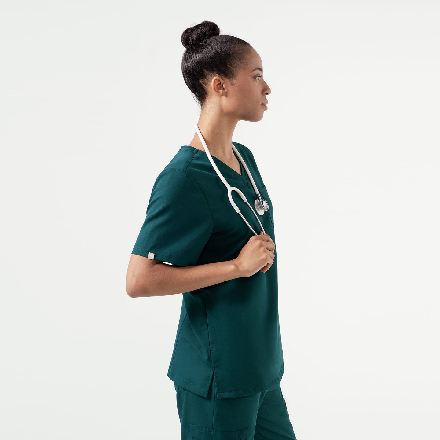 A person wearing a Pine Green scrub set stands in profile with a stethoscope around their neck, showcasing the clean V-neck and streamlined fit of the top.