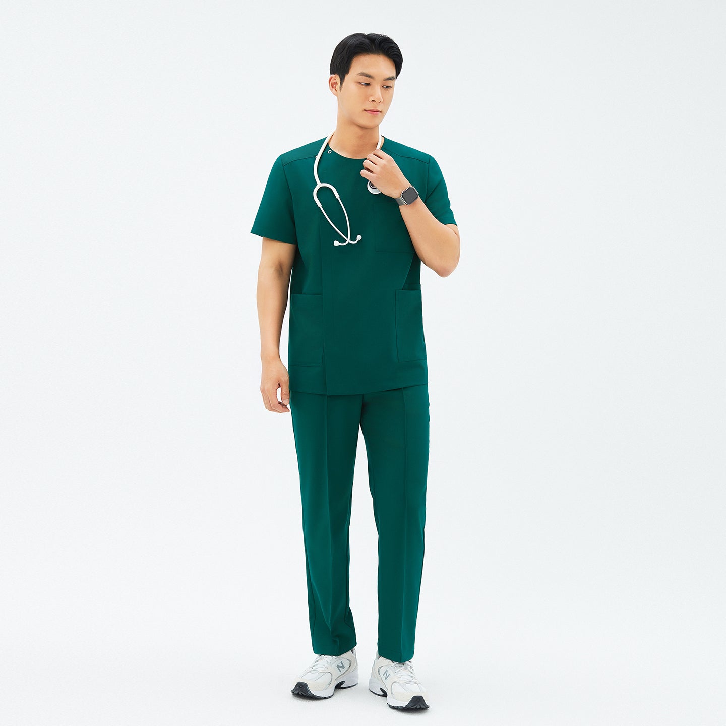 Image of Full-length shot of a male model in TUL Men’s Premium Banding Pants (Pine Green) and a matching scrub top, holding a stethoscope around his neck. He wears white athletic shoes and a smartwatch, creating a cohesive medical uniform.