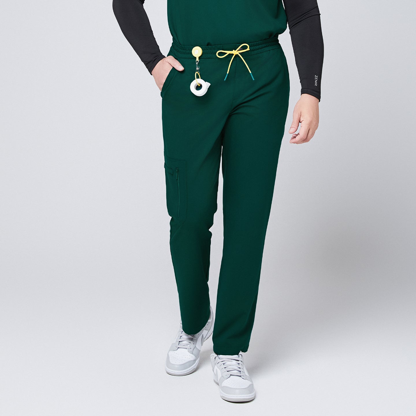 Image of Male model walking forward, showcasing pine green straight-leg scrub pants with a yellow drawstring and zipper pockets, paired with white sneakers and a black undershirt,Pine Green