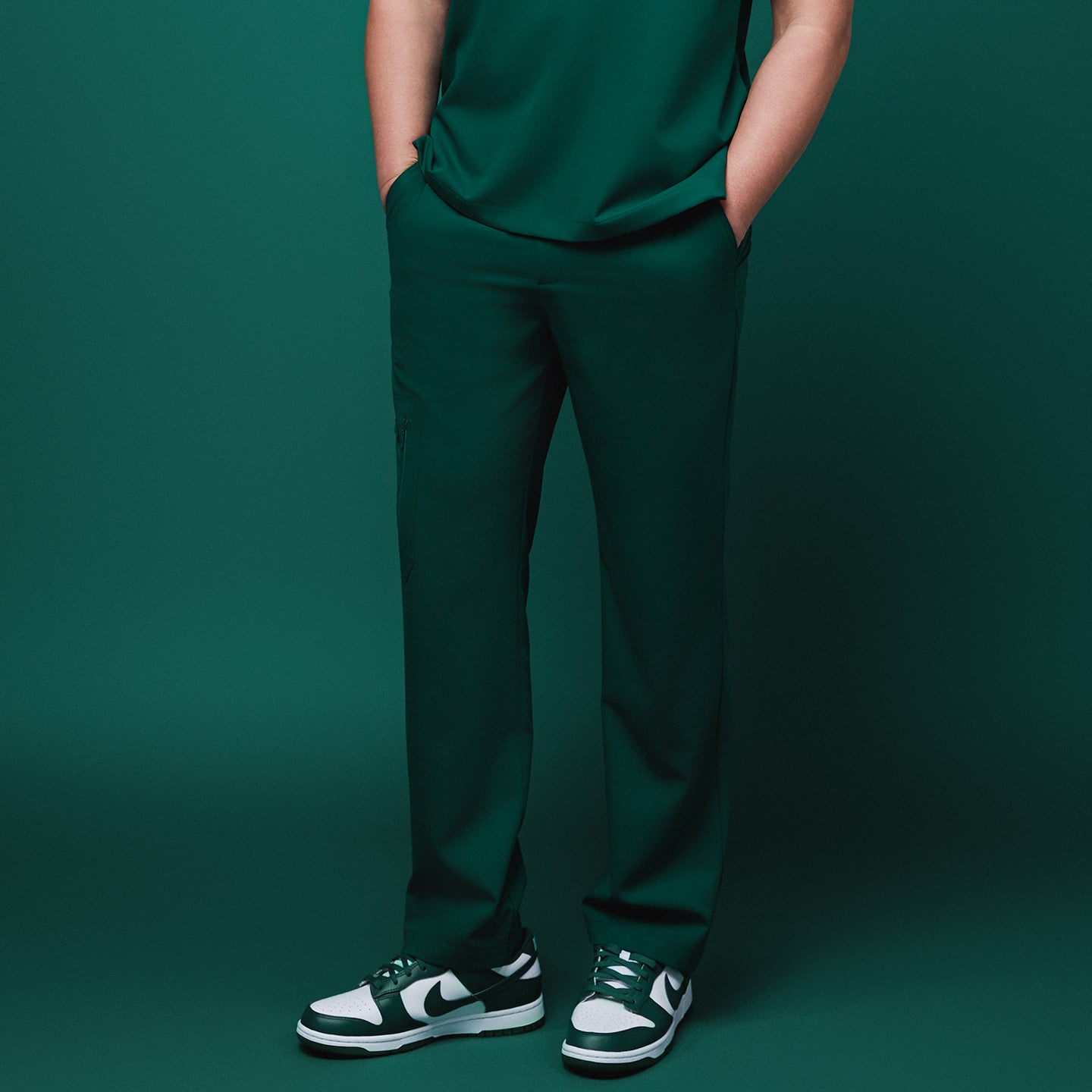 Male model wearing pine green straight-leg scrub pants with zipper pockets, hands in pockets, paired with green and white sneakers, against a matching green background,Pine Green