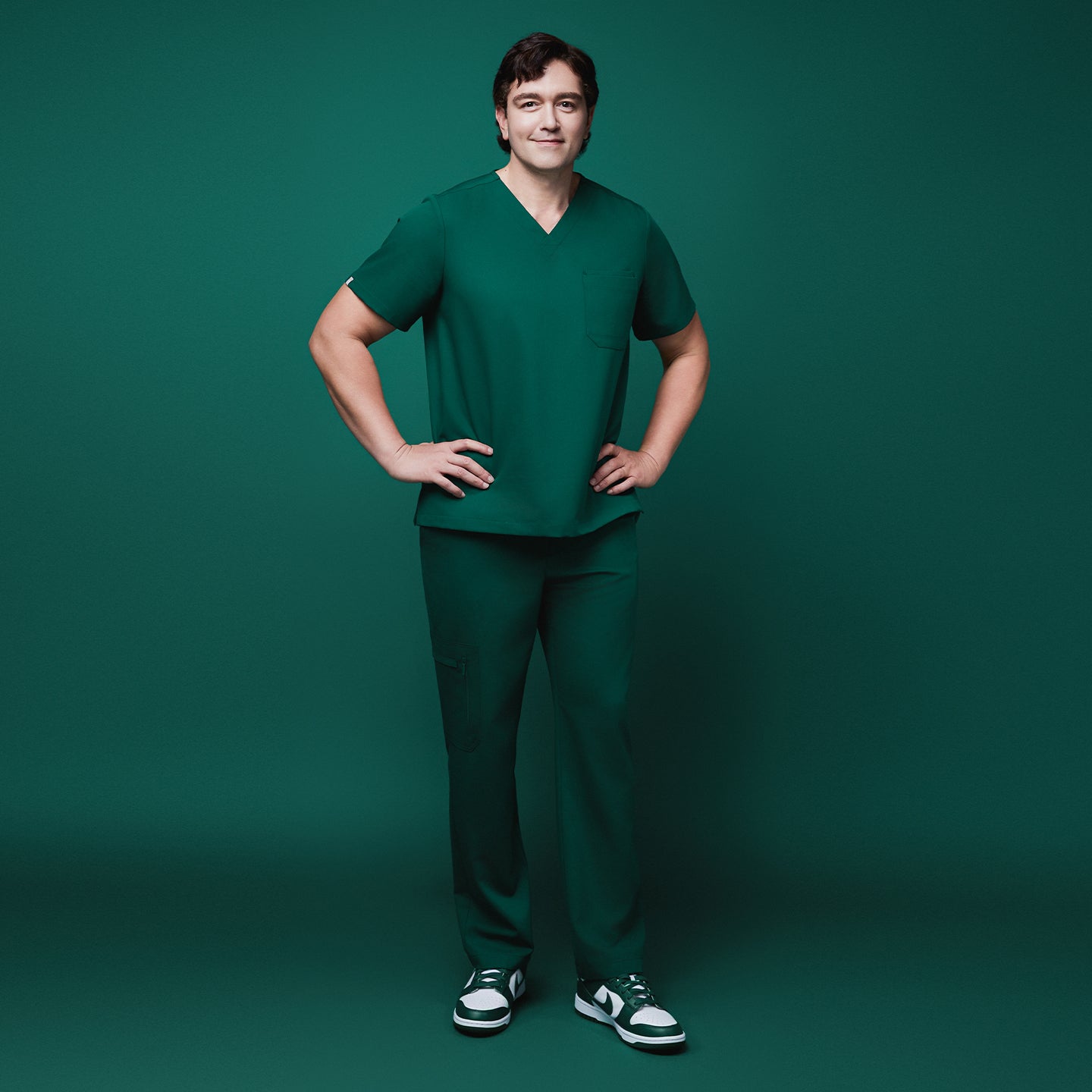 Male model standing confidently in full-body view, wearing pine green scrubs with straight-leg pants and zipper pockets, styled with green and white sneakers, set against a matching green background,Pine Green