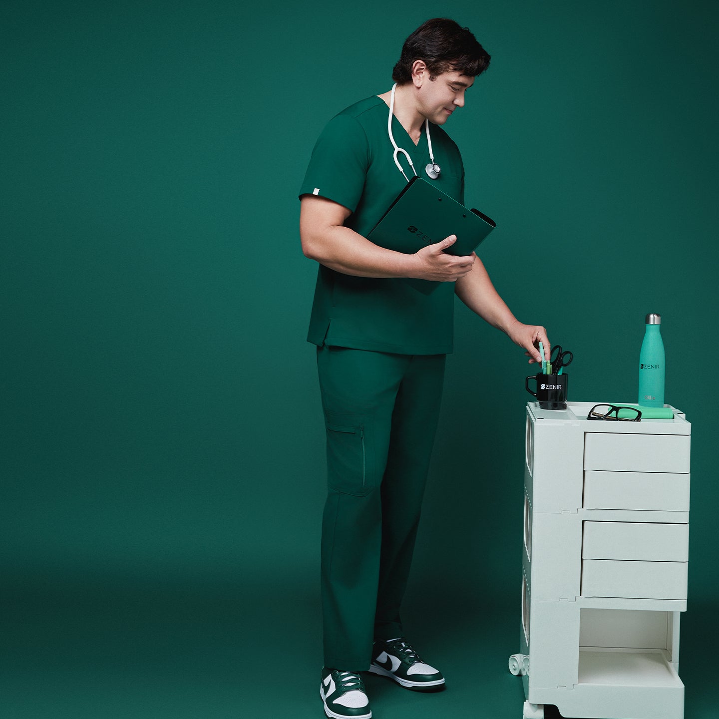 Image of Male model wearing pine green scrub set with straight-leg zipper pocket pants, holding a clipboard and interacting with a rolling cart featuring branded items, against a green background,Pine Green