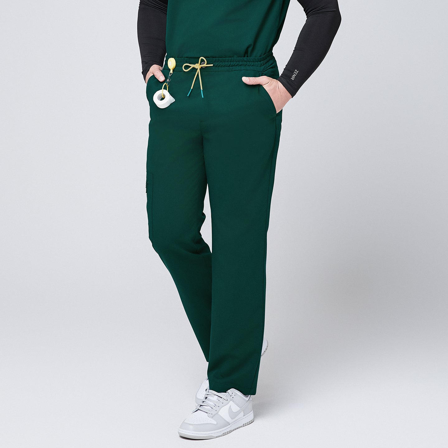Male model showcasing pine green straight-leg scrub pants with a yellow drawstring and zipper pockets, paired with white sneakers and a black undershirt, standing in a casual pose,Pine Green