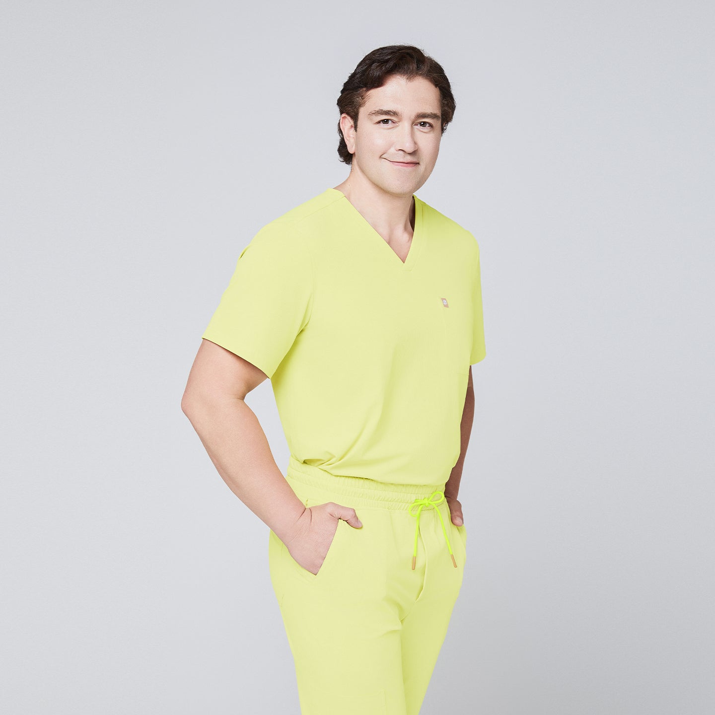 Image of Male model wearing a pineapple yellow V-neck single-pocket scrub top and matching pants, smiling with hands in pockets,Pineapple Yellow