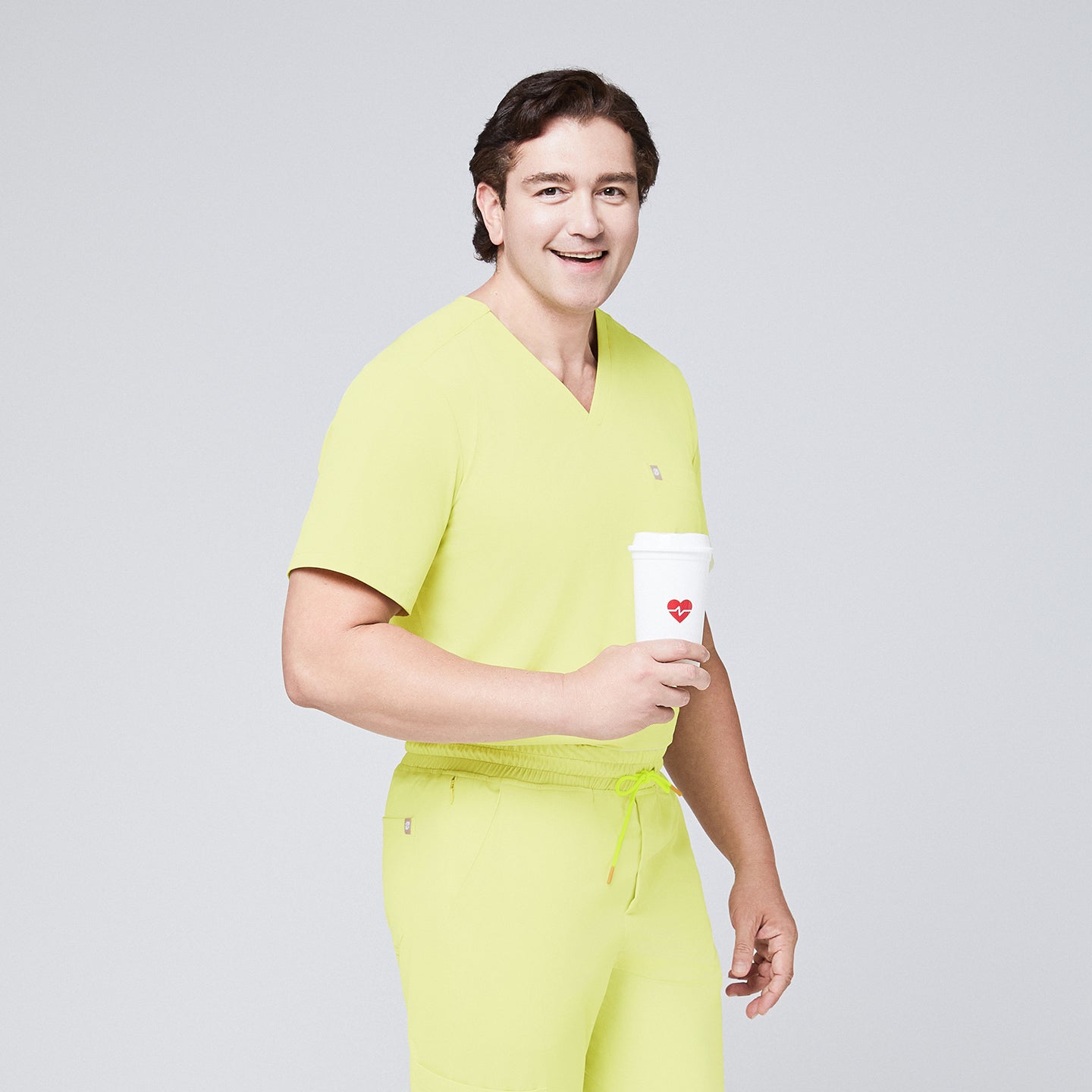 Image of Male model in a pineapple yellow V-neck single-pocket scrub top, holding a coffee cup with a heart design and smiling,Pineapple Yellow