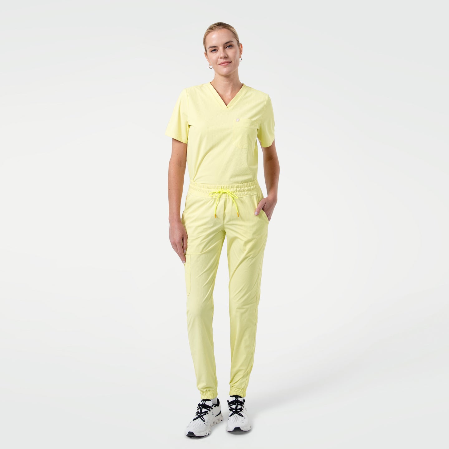 Standing with one hand in the pocket, the model wears a Pineapple Yellow V-neck scrub top with short sleeves and a chest pocket, paired with matching jogger-style scrub pants and white sneakers against a clean light background.